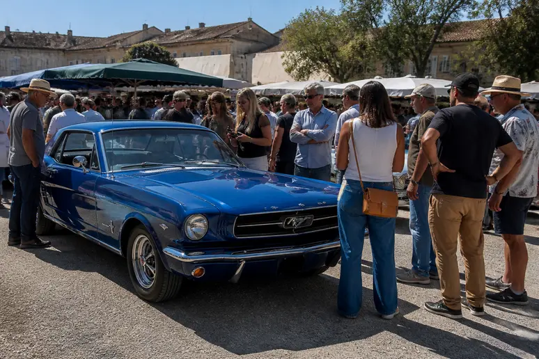 Des passionnés de voitures anciennes examinent une Ford Mustang bleue vintage lors d'un marché en plein air, sous un ciel ensoleillé.
