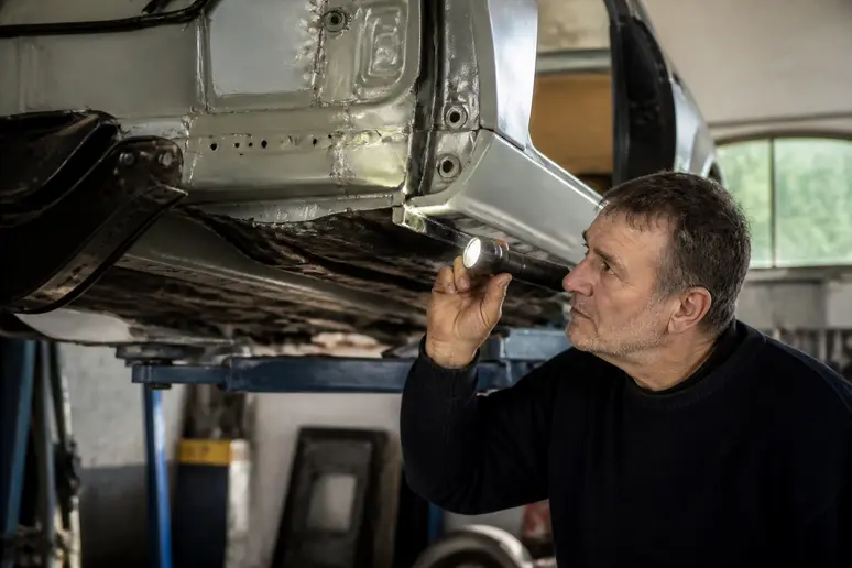 Un homme d'âge mûr examine méticuleusement le châssis d'une Peugeot 504 coupé dans un garage bien éclairé, soulignant l'importance de l'état de la carrosserie pour la valeur.
