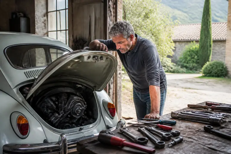 Homme français inspectant le moteur d'une Coccinelle classique dans un garage ensoleillé, outils à proximité.